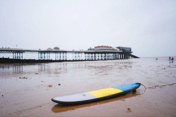 An image of Cromer Pier extending into the sea with a cloudy sky overhead and a colorful surfboard lying on the wet sand in the foreground.