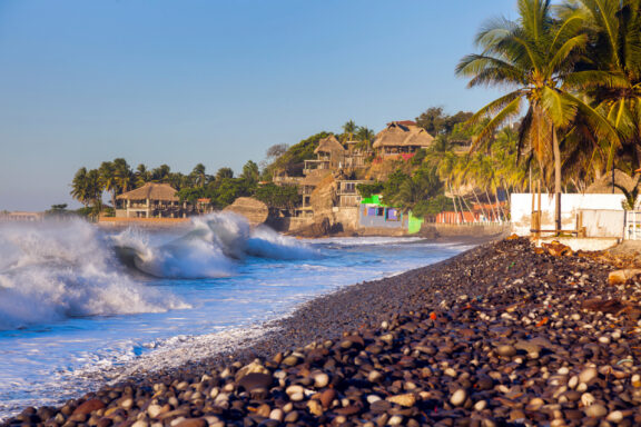 El Tunco Beach in El Salvador displays waves hitting a rocky shore, palm trees, buildings, under a clear sky.