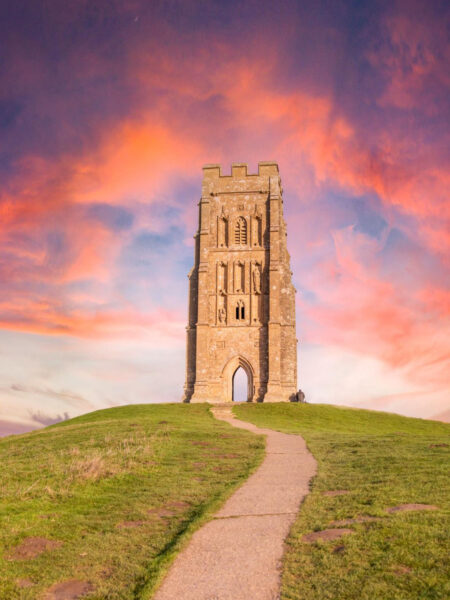 A stone tower stands atop a hill under a sky with pink and orange hues, likely Glastonbury Tor in England. A path leads up to the tower.