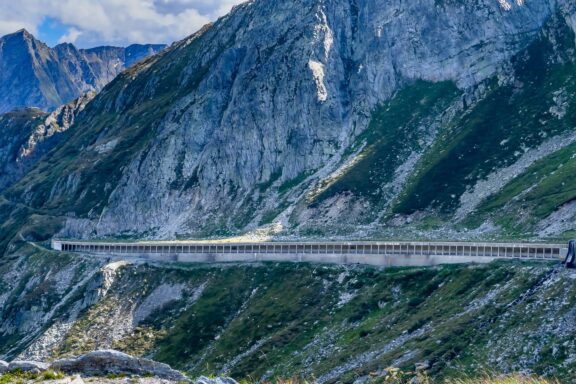 A road tunnel entrance, possibly the Gotthard Road Tunnel in Switzerland, is built into a rocky, sparsely vegetated mountain landscape.