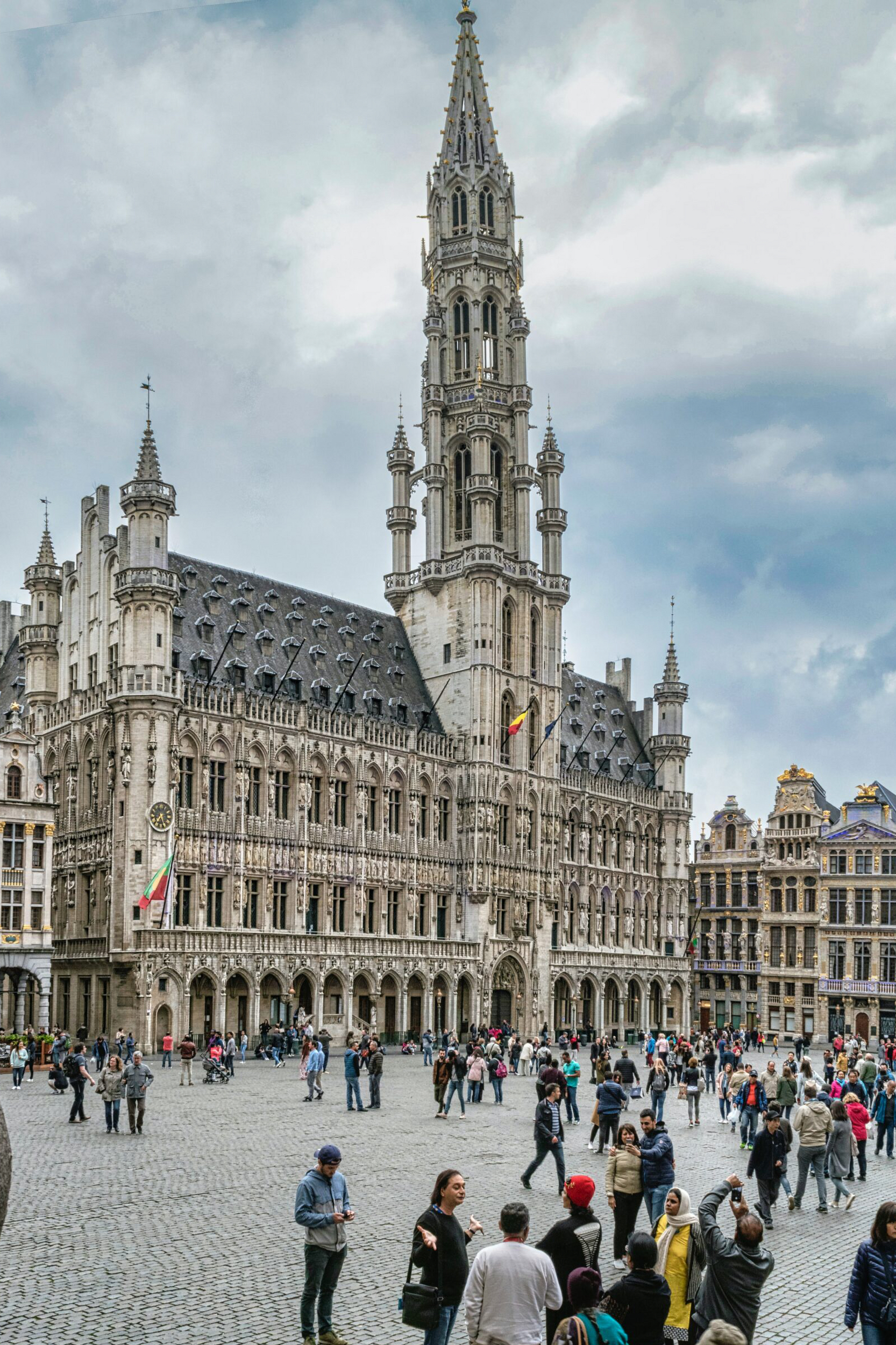 A vertical image showing a crowd of people at the Grand Place with the Gothic architecture of the Town Hall in Brussels, Belgium, under a cloudy sky.