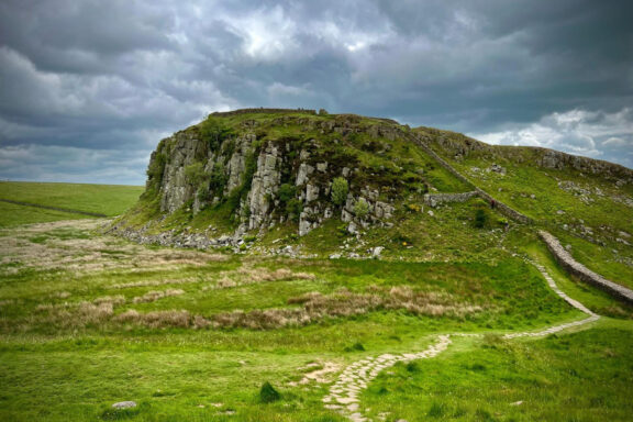 A picturesque scene of Hadrian's Wall in Northumberland with a grassy rock formation and footpath under a cloudy sky.