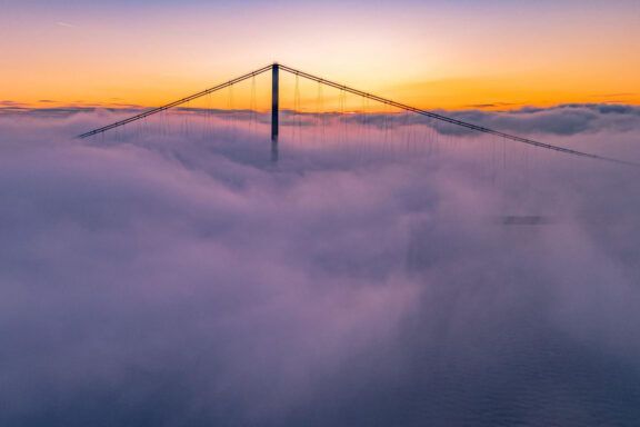 Aerial shot of UK's Humber Bridge in Hessle, partly hidden by clouds at sunrise/sunset, with a warm-colored sky.