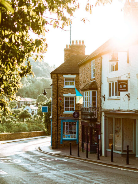 A quaint street scene in Ironbridge town with sunlight filtering through trees, showcasing historic buildings with a clear sky.