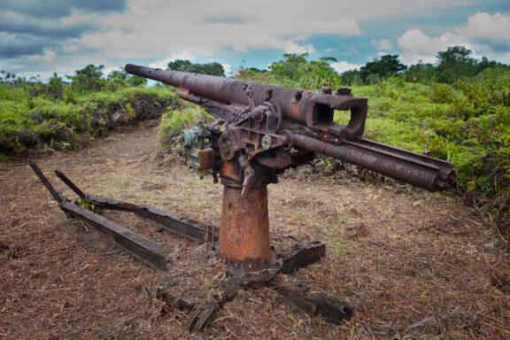 A rusted Japanese anti-aircraft gun in a grassy area with trees in the background, located in Nefo Cave, Micronesia.