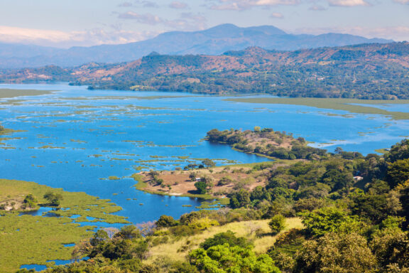 A scenic view of Lake Suchitlán in El Salvador, featuring blue waters with green vegetation and surrounding mountains under a clear sky.