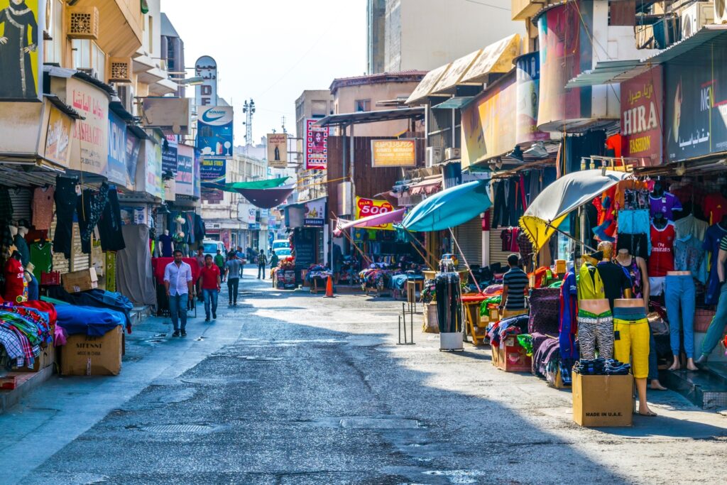 A vibrant street scene at Manama Souq, Bahrain, featuring stalls selling colorful goods and bustling with people.