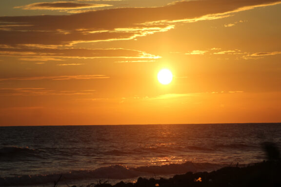 Sunset at Mizata Beach, El Salvador features a low sun, orange-yellow sky, gentle waves, and visible rocks.