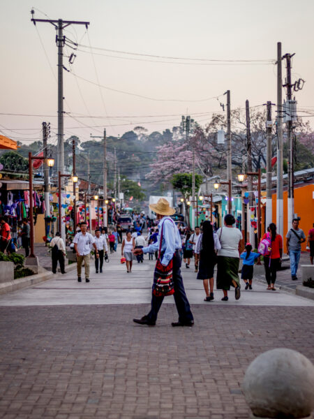 A person in traditional attire strolls a busy, colorful street in Nahuizalco, El Salvador, with hills in the backdrop.