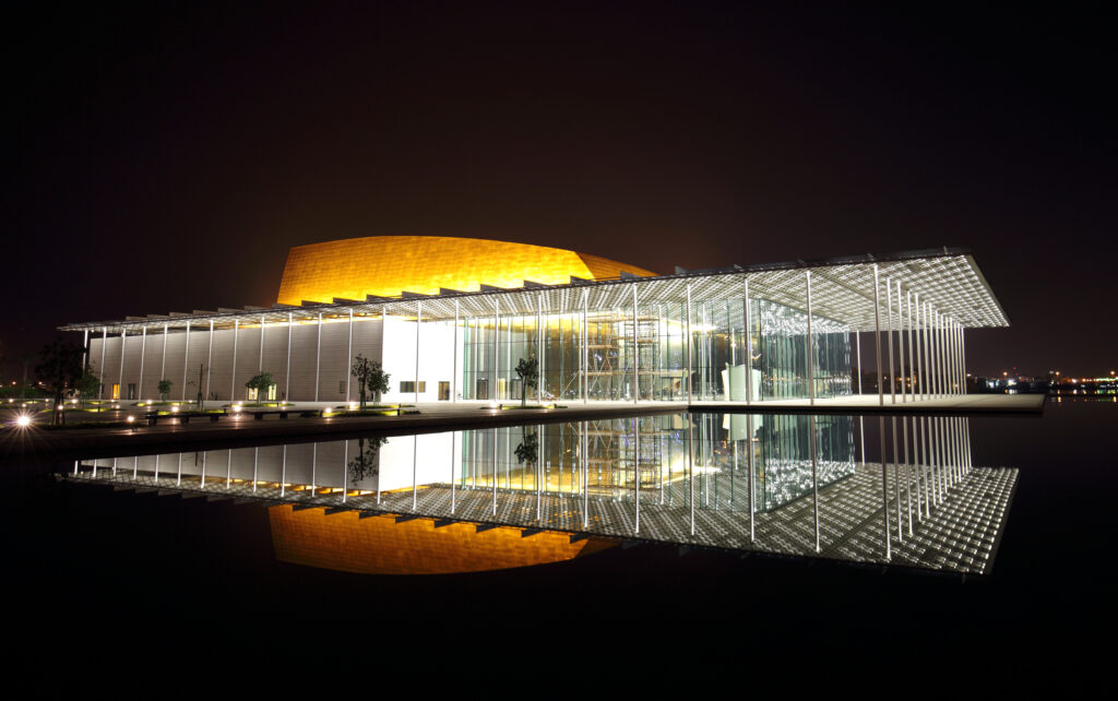 A night view of the National Theater of Bahrain, featuring its illuminated facade and reflection on the water surface.