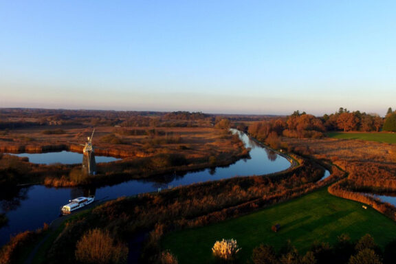 Sunset aerial view of Norfolk Broads with a winding river, windmill, boat, greenery, and clear sky.