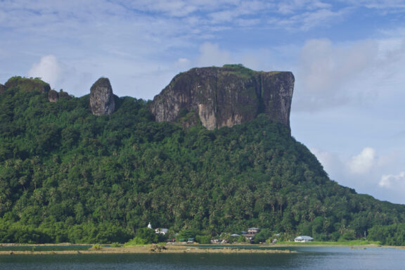 A lush green landscape with Sokehs Rock, a prominent cliff, on the island of Pohnpei in Micronesia, under a partly cloudy sky.