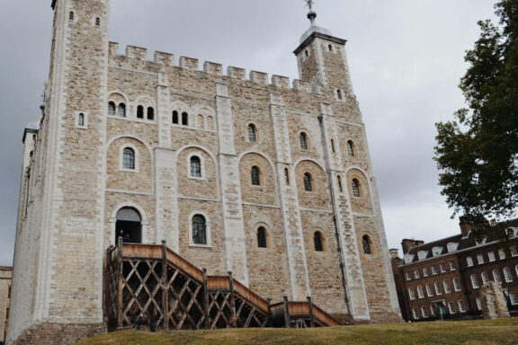 An image of the Tower of London, featuring the historic stone fortress with a wooden gate, under a cloudy sky.