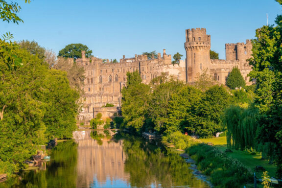 A picturesque view of Warwick Castle with its medieval towers and ramparts, reflected in the tranquil waters of a river surrounded by lush greenery.