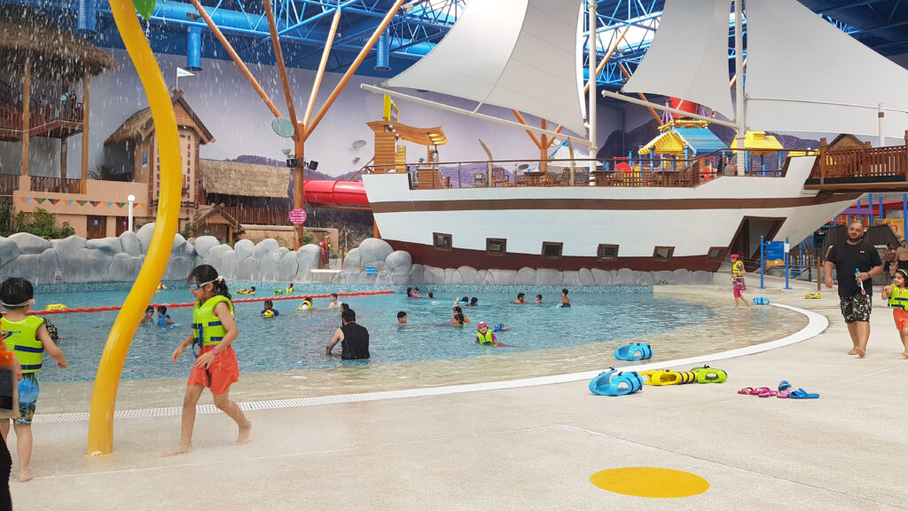 Indoor water park with visitors swimming and walking around, featuring a large pool, a ship-themed structure, and a colorful water play area.