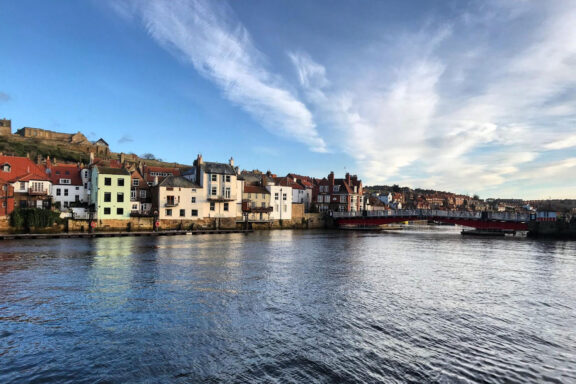 A scenic view of Whitby, UK, featuring colorful buildings along the waterfront with a clear sky above and a calm river in the foreground.