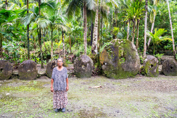 A person standing in front of large stone discs surrounded by tropical vegetation on Yap Island, Micronesia.