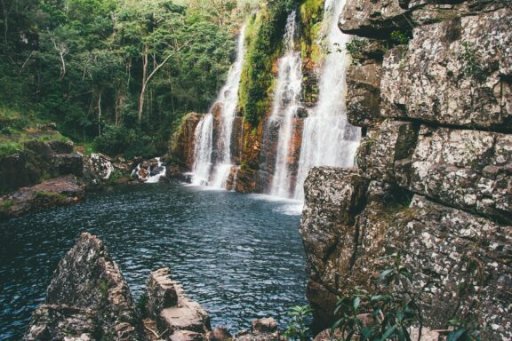 A peaceful waterfall with many cascades flows into a tranquil pool at Almécegas I, Brazil, surrounded by lush greenery and rocks.