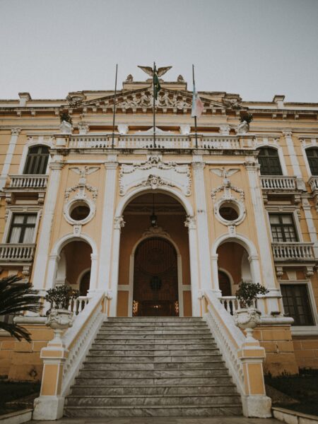 A photo displays the baroque architecture of Brazil's Anchieta Palace, with a grand staircase, arched entrance, and ornate columns.