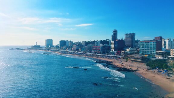 A picturesque coastal view of Barra, Salvador, Brazil, with a blue sky, calm ocean, sandy beach, cityscape, and distant lighthouse.