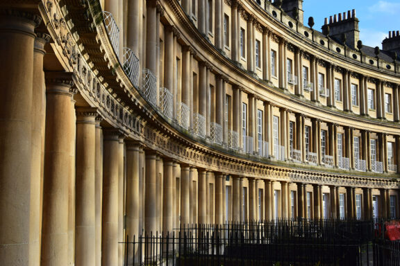 An image showing a curved row of Georgian architecture with columns in Bath, England, bathed in warm sunlight.