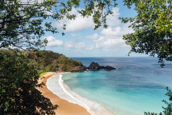 A secluded beach with golden sand in Fernando de Noronha, surrounded by lush greenery and clear blue waters.