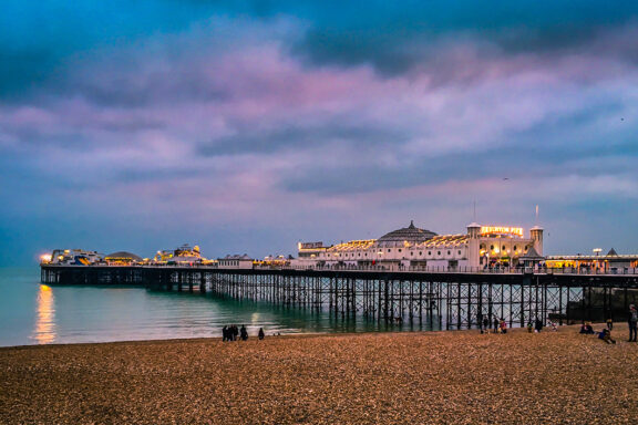 A scenic view of Brighton Pier at dusk with a colorful sky, the pier illuminated by lights, and people on the pebble beach in the foreground.