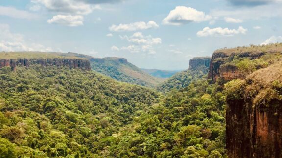 A panoramic view of Chapada dos Guimarães in Brazil, showcasing lush greenery, towering cliffs, and a vast canyon under a partly cloudy sky.