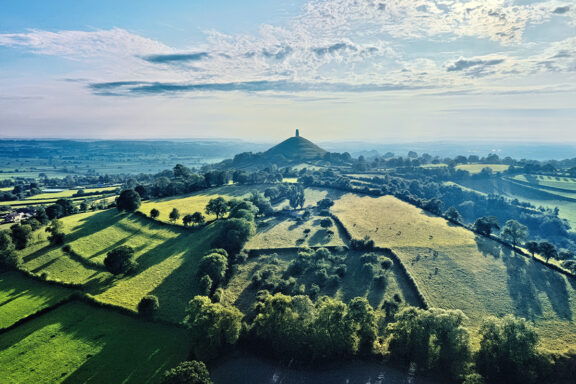 Aerial view of green fields, hedgerows near Glastonbury, with a distant hilltop tower under a partly cloudy sky.