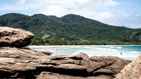 A picturesque view of Ilha Grande, Brazil, with a sandy beach, turquoise waters, green mountains, and a rocky foreground.