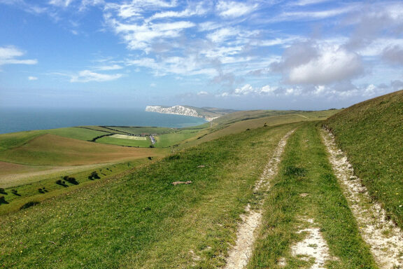 A picturesque view of a grassy trail through hills, with a distant ocean and cliff, under a partly cloudy sky on the Isle of Wight.