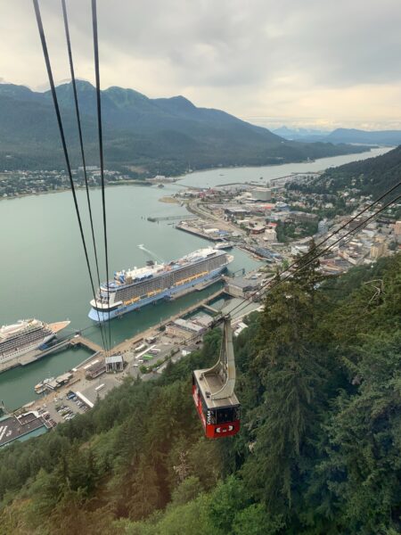 Aerial view of Juneau, Alaska, showing a red cable car in the foreground with a cruise ship docked in the harbor and mountains in the background.