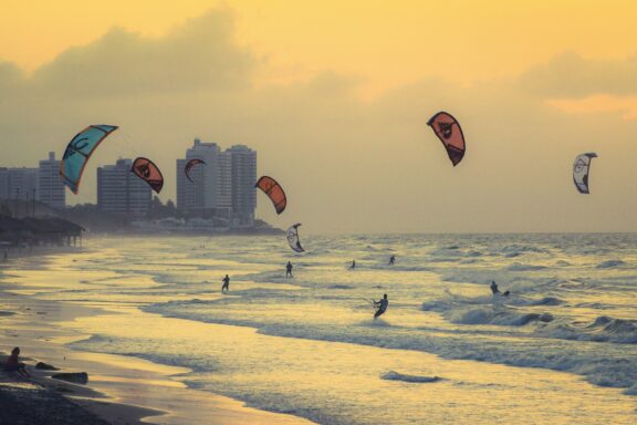 A sunset view of a beach in Brazil with multiple kitesurfers on the water and buildings in the background.