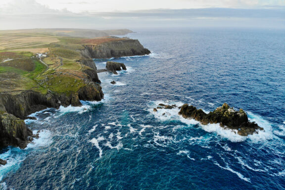 Aerial view of Lands End, featuring rugged cliffs along the coastline with waves crashing against the rocks, under a partly cloudy sky.