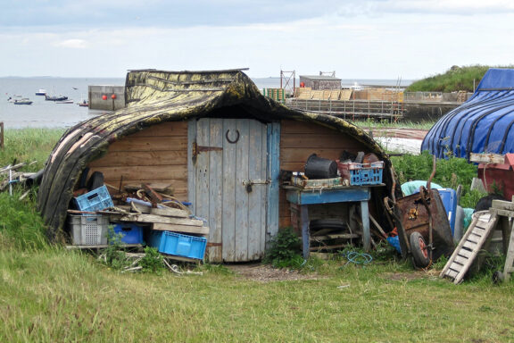 A rustic shed, possibly an upturned boat, sits among debris in Lindisfarne, with a water body and boats in the background.