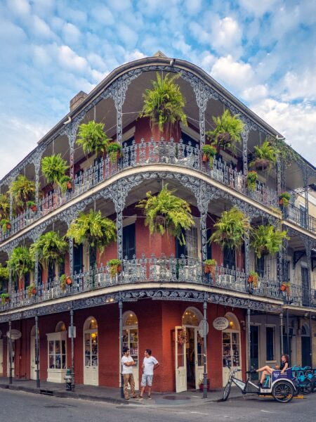 A traditional New Orleans building with iron balconies and plants, under a cloudy sky. People, including a rickshaw cyclist, are on the sidewalk.