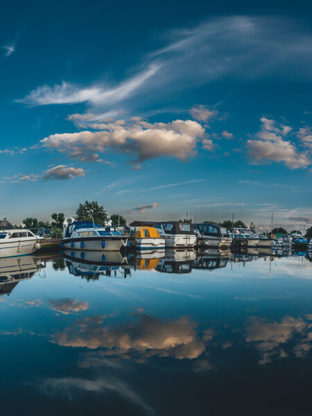 A serene image of the Norfolk Broads showing boats moored on calm water with reflections of the boats and a blue sky with fluffy clouds.