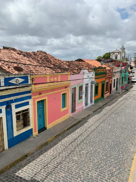 A street view of colorful colonial buildings in Olinda, Pernambuco, Brazil, with cobblestone pavement and a cloudy sky.