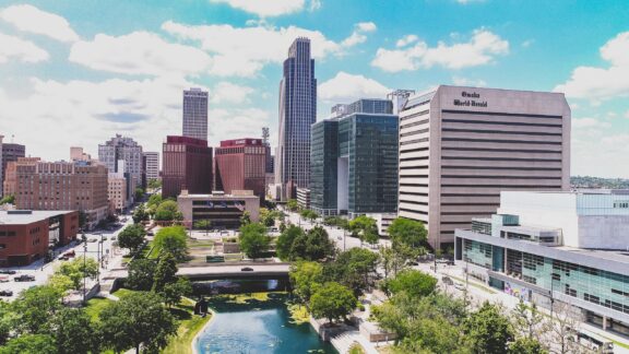 Aerial view of downtown Omaha, Nebraska, featuring modern buildings, a river, and greenery under a partly cloudy sky.