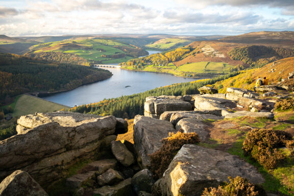 A scenic view of the Peak District with a lake surrounded by forested hills and rugged terrain, under a partly cloudy sky.