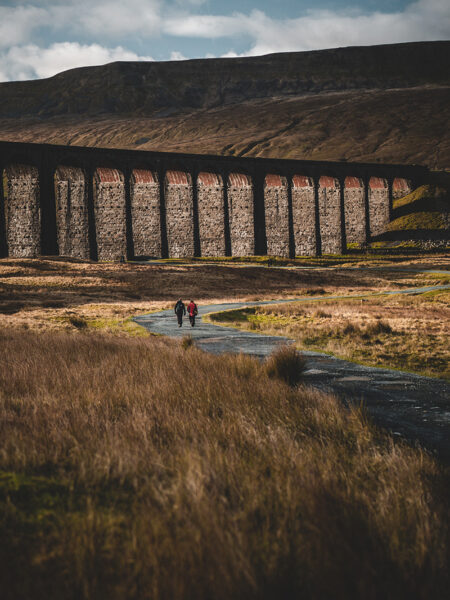 Two people walk on a path through a field with the Ribblehead Viaduct, a large stone railway bridge, in the background.