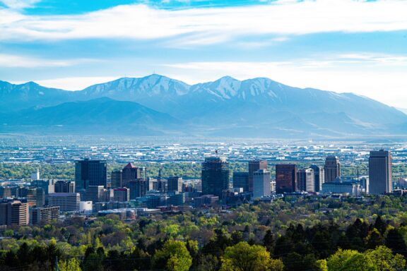 A panoramic view of Salt Lake City with a skyline of buildings against a backdrop of mountains and a clear blue sky.