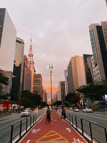 A dusk street view in São Paulo, Brazil, featuring a bike lane, pedestrians, tall buildings, and a colorful sky.