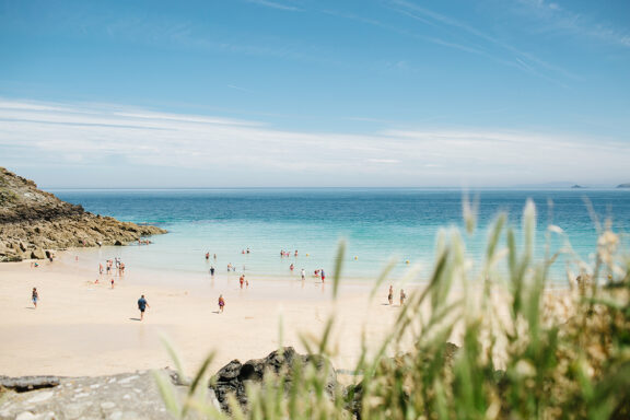 People enjoy sunny weather at a scenic St Ives beach, with clear skies, turquoise waters, and visible grass and rocks.