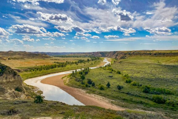 A scenic view of Theodore Roosevelt National Park in North Dakota, featuring rolling hills, a winding river, and a partly cloudy sky.