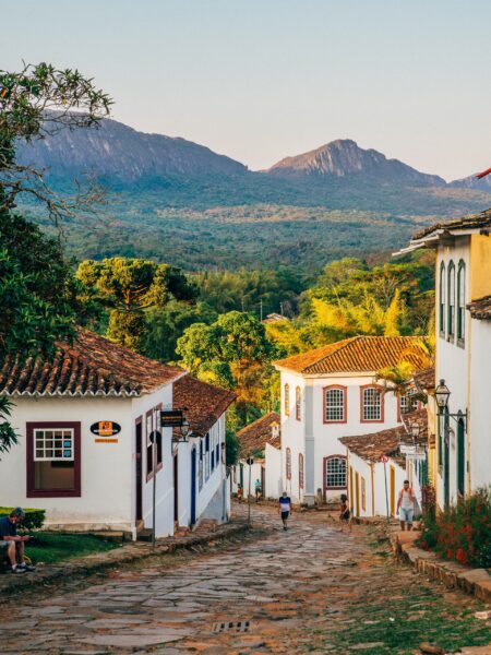 A scenic street in Tiradentes, Brazil features colonial buildings, cobblestone paths, and is surrounded by green mountains.