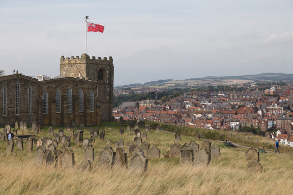 A scene of Whitby features an ancient church's ruins, a graveyard, a red flag, and a dense housing array in the distance.