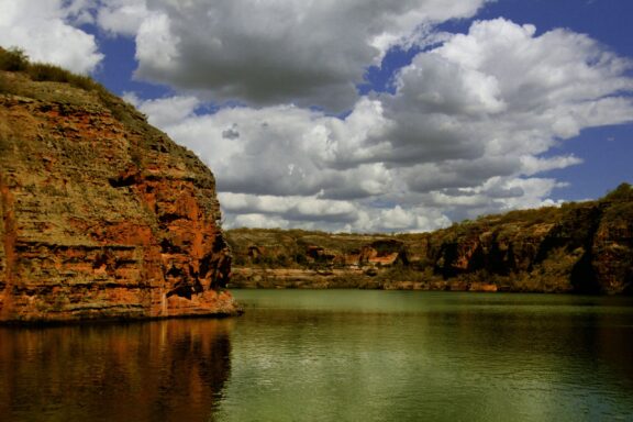 A view of Xingó Canyon with red rock formations beside calm green waters under a cloudy sky in Sergipe, Brazil.