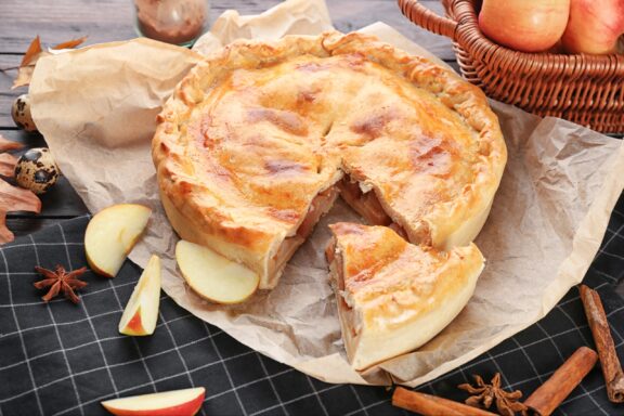 A new apple pie with a slice taken out, on a dark surface with apples, cinnamon sticks, and spices around it.