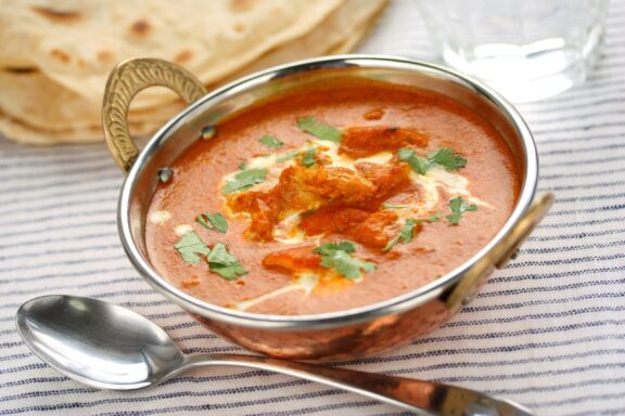 A bowl of butter chicken, an Indian dish, garnished with herbs, with flatbread and a spoon on the side.